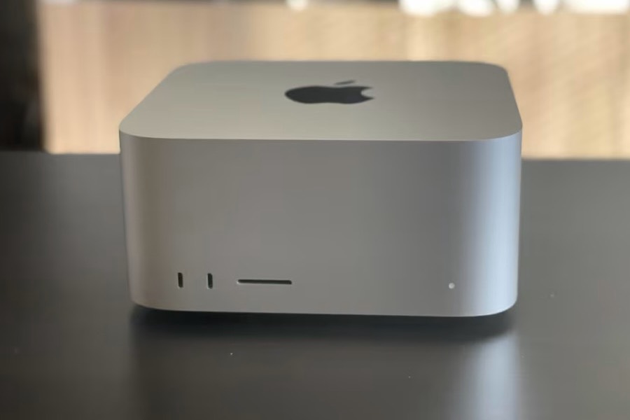 A silver Mac Studio sits head-on on a dark desk &mdash; two USB-C ports, a card slot and a small white power light along its front edge. The room behind it is warm, out of focus and unreadable.